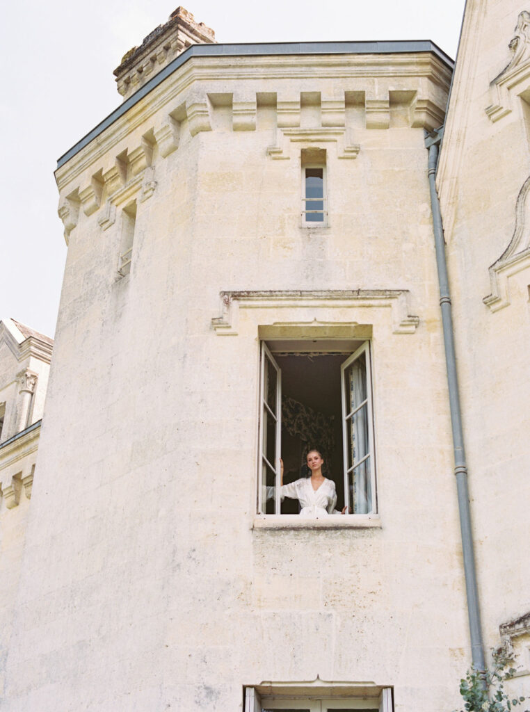 Woman in a white dress leans on the window sill ofChâteau Le Petit Verdus