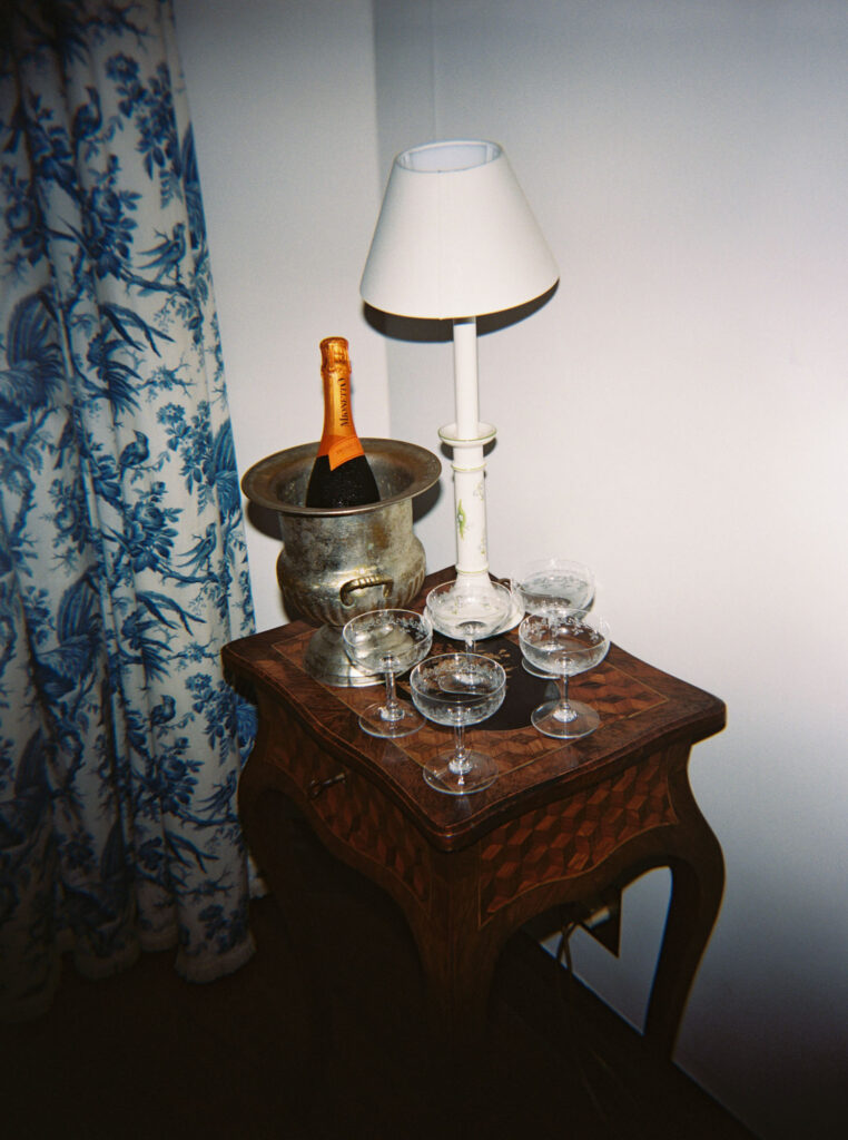 Wooden side table holds a champagne bottle in a vintage ice bucket, a white table lamp, and four empty champagne glasses