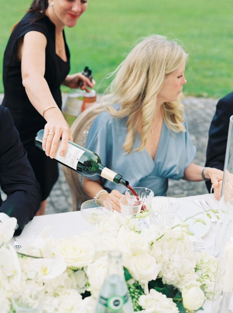 Person in a black dress pouring red wine into a glass and a woman seated next to them at a table decorated with white flowers, next to an open bottle of sparkling water