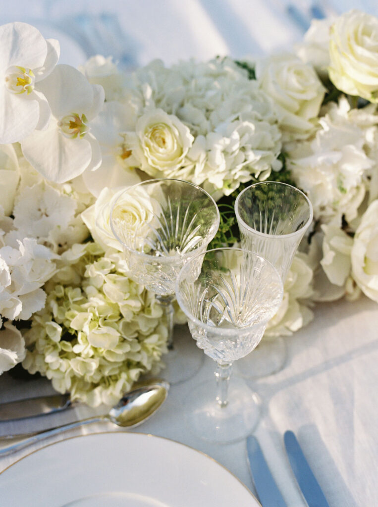 Table setting featuring crystal glassware and a lush floral arrangement set against a white tablecloth with a hint of polished silverware and a white plate with a gold rim