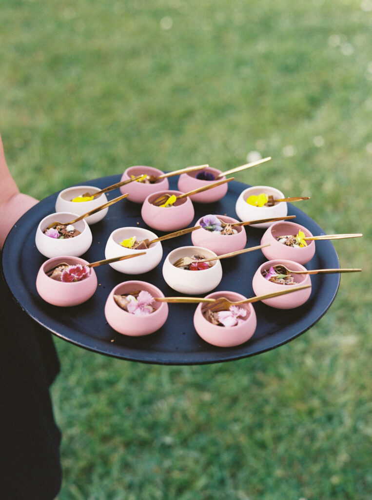 Person holding a round black tray with several small, colorful bowls filled with garnished appetizers on wooden skewers