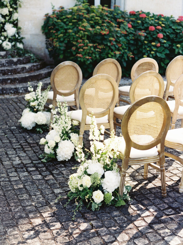 Outdoor wedding ceremony setup with rustic wooden chairs arranged in rows on a cobblestone path adorned with arrangements of white flowers