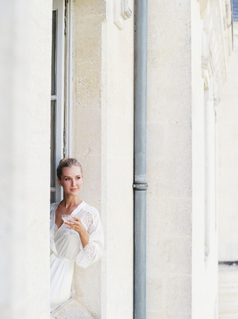 Bride in a white lace-trimmed robe leans out  Château Le Petit Verdus' window, smiling and holding a glass