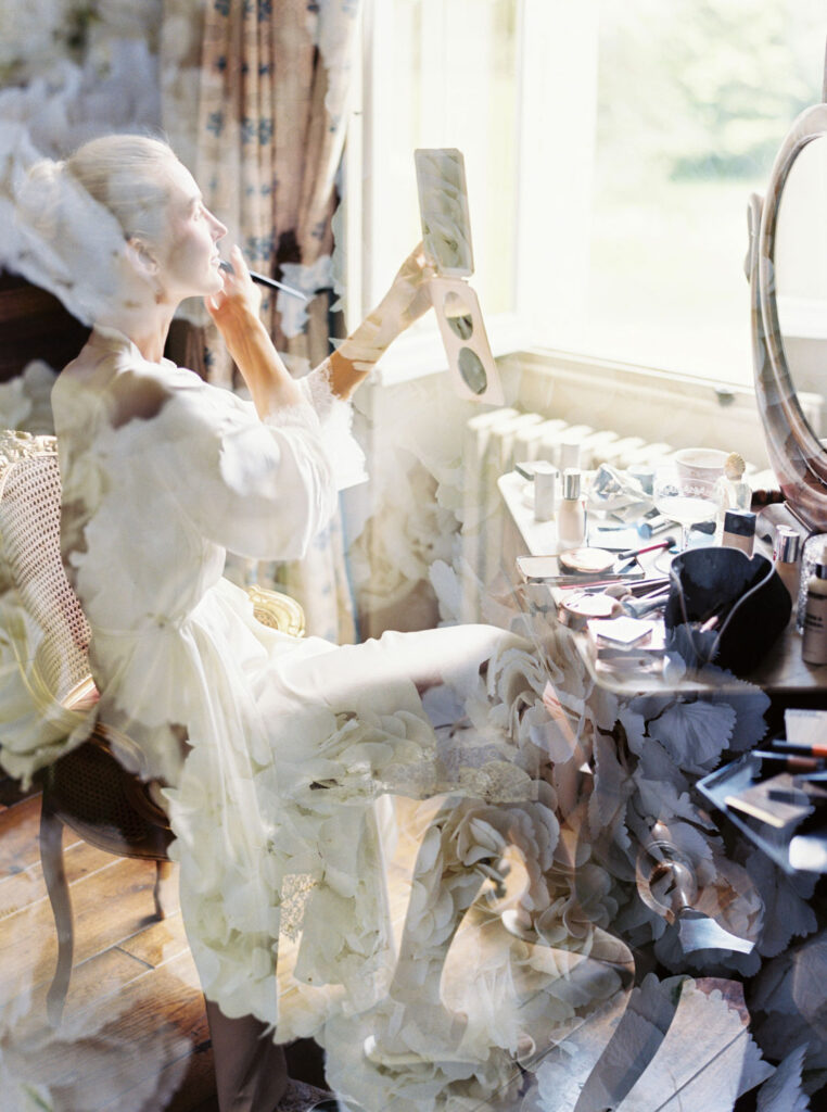 Bride in her white robe sits at a vanity table, applying makeup while looking into a handheld mirror