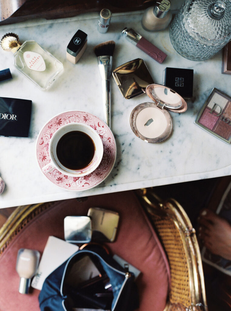 A flat lay photo of a cluttered vanity that has a marble surface with a red-patterned cup of black coffee, makeup items including foundation, powders, a brush, and a Dior and Givenchy case