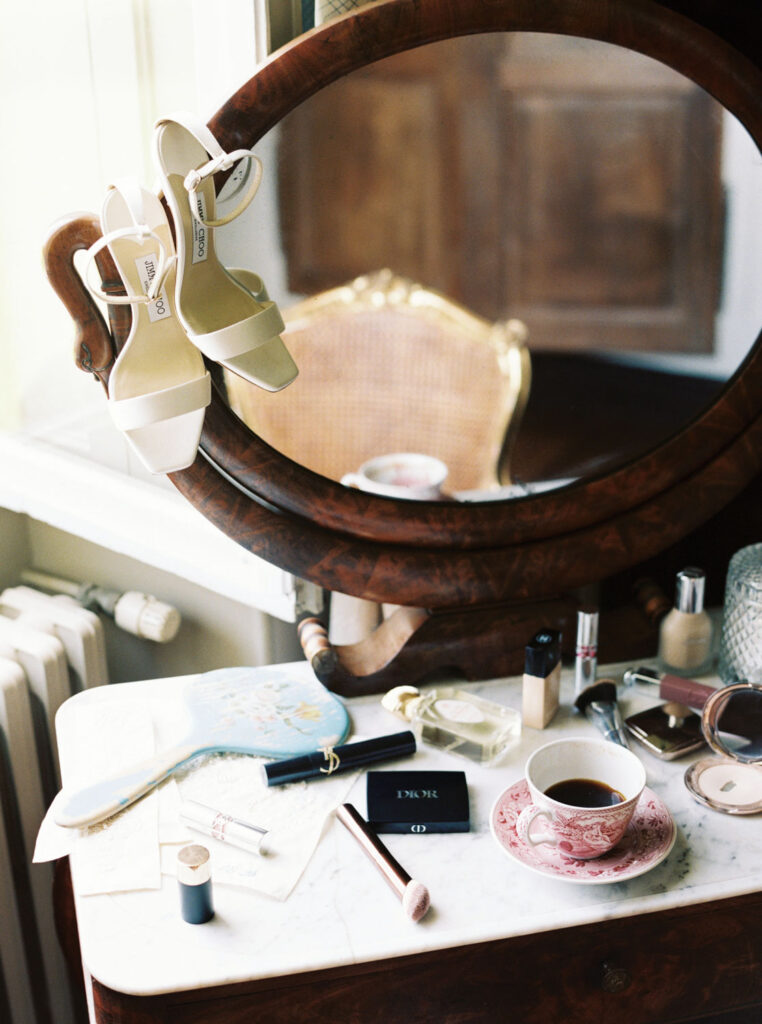 Vintage vanity table with a round mirror displays white heeled sandals hanging on its frame and makeup items, perfume bottles, a hairbrush, and an ornate teacup filled with coffee