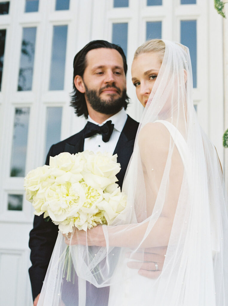 Bride, holding a bouquet of white roses, wearing a white gown and a sheer veil while looking to the side and groom, in a black tuxedo, stands beside her, gazing at her with a smile