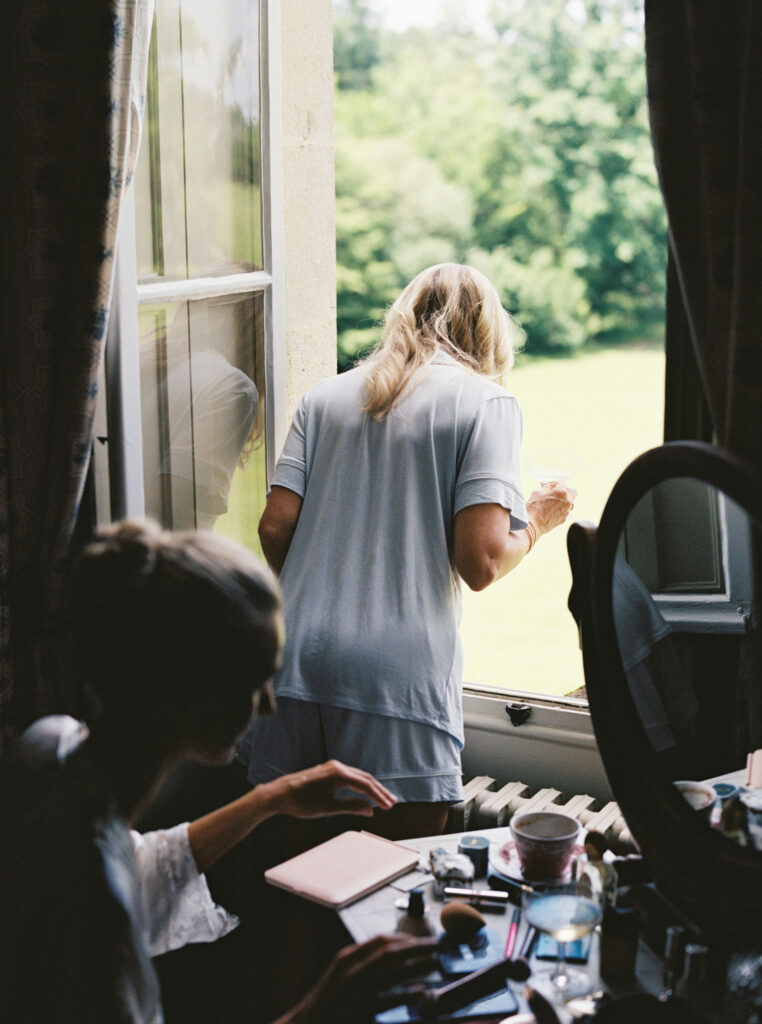 Bride leans out of an open window, holding a drink and another person sitting beside a desk cluttered with makeup items and a large round mirror, with green foliage visible outside