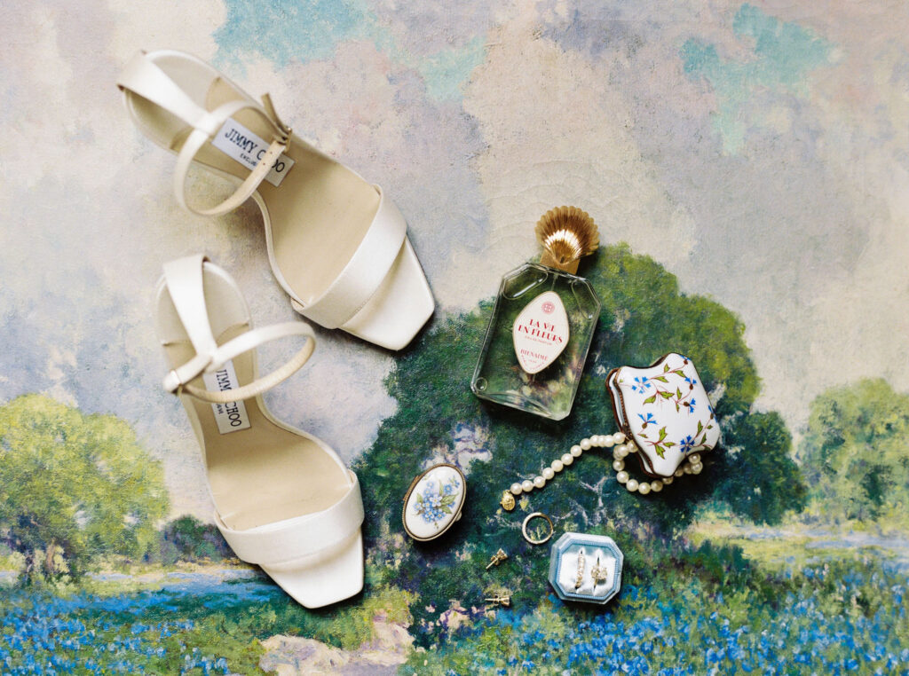 Flat lay of wedding items on a painted floral backdrop includes white heeled shoes, a bottle of perfume, a small floral tin with pearls, two small oval trinket boxes, and a ring box with gold earrings