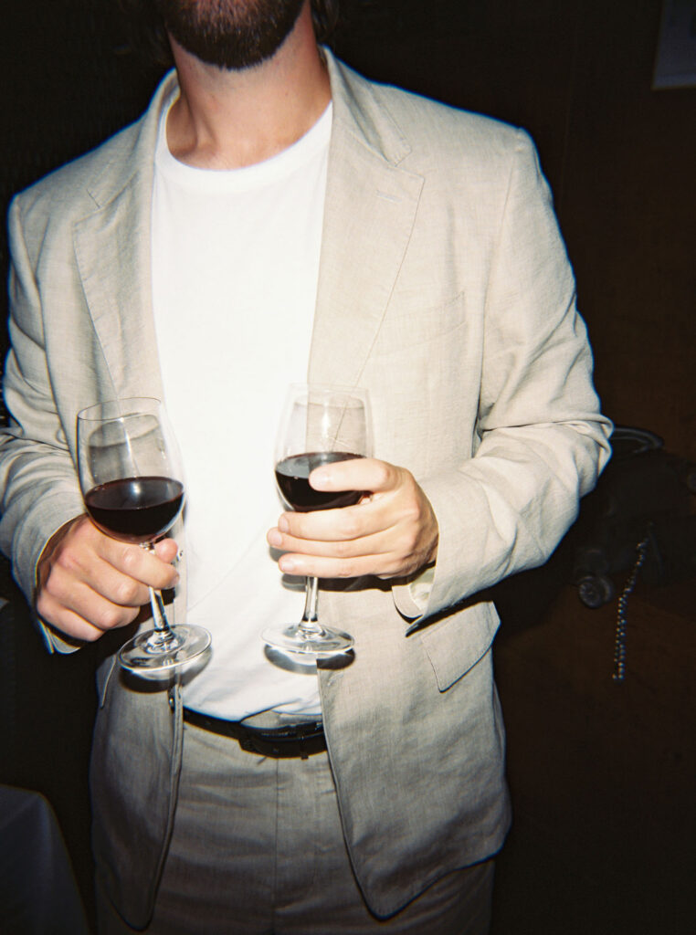 Person wearing a cream-colored suit and white t-shirt is holding two glasses of red wine