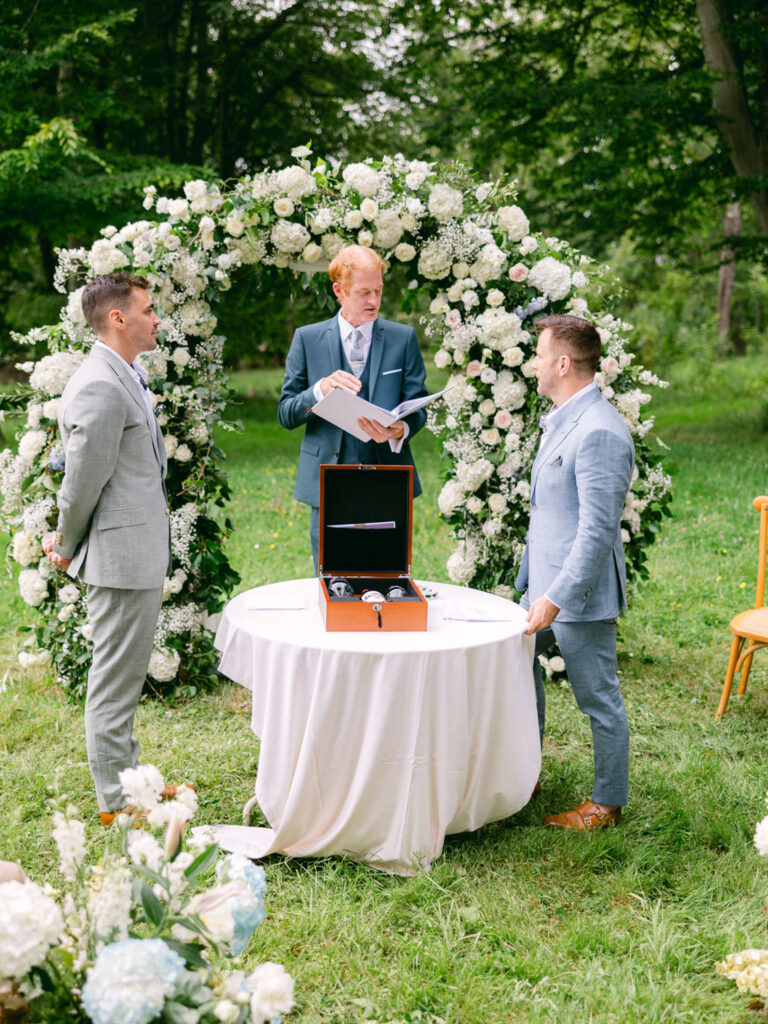 Kevin and Steven stand together at their outdoor wedding ceremony in France, with a floral arch and an officiant present