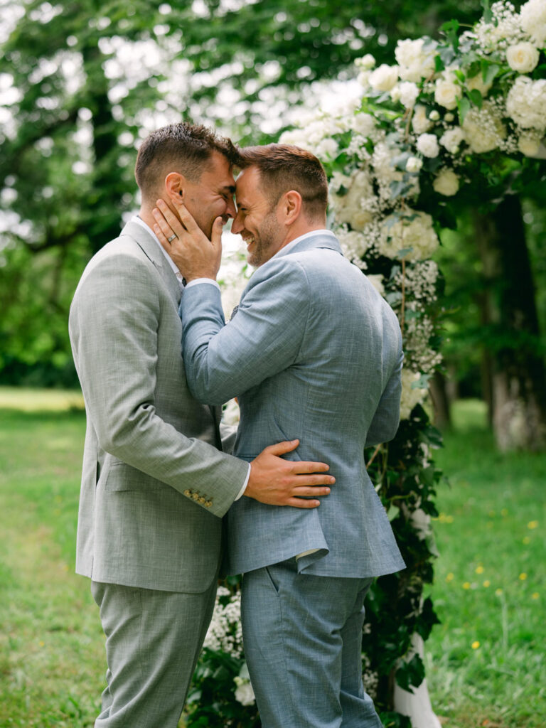 Kevin and Steven share an emotional embrace during their wedding ceremony in France, framed by a beautiful floral arch