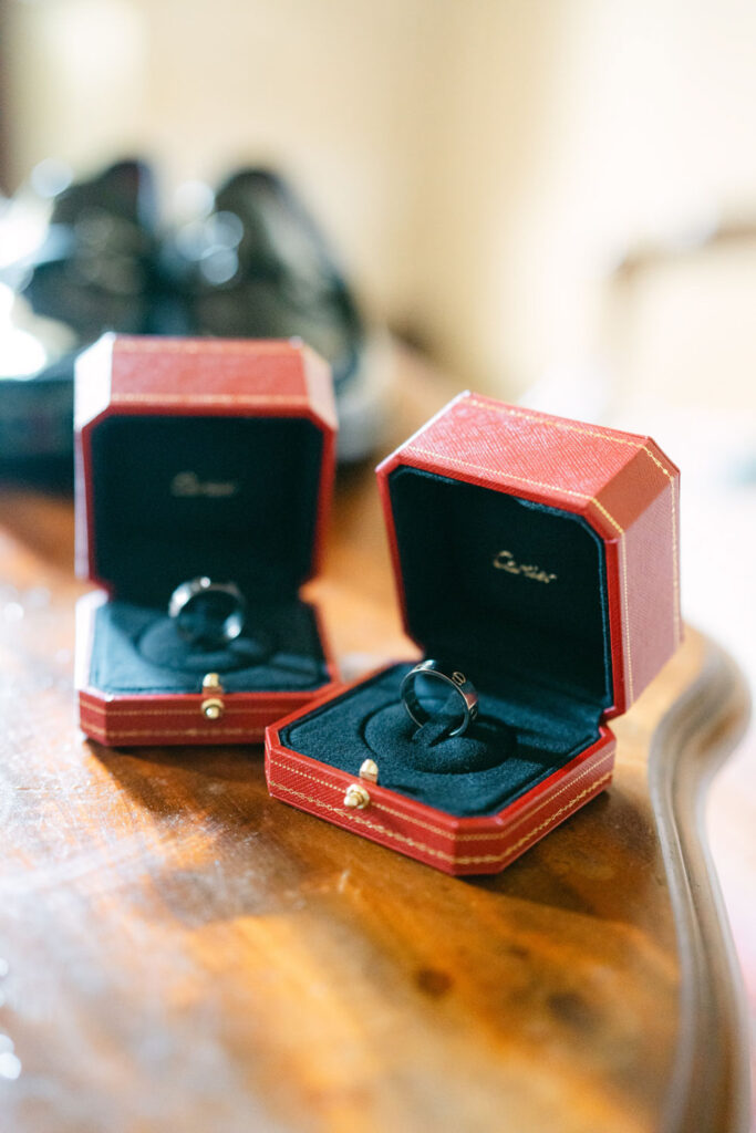 Two Cartier red ring boxes open, each displaying a black ring, placed on a wooden surface and a pair of black shoes is slightly blurred in the background