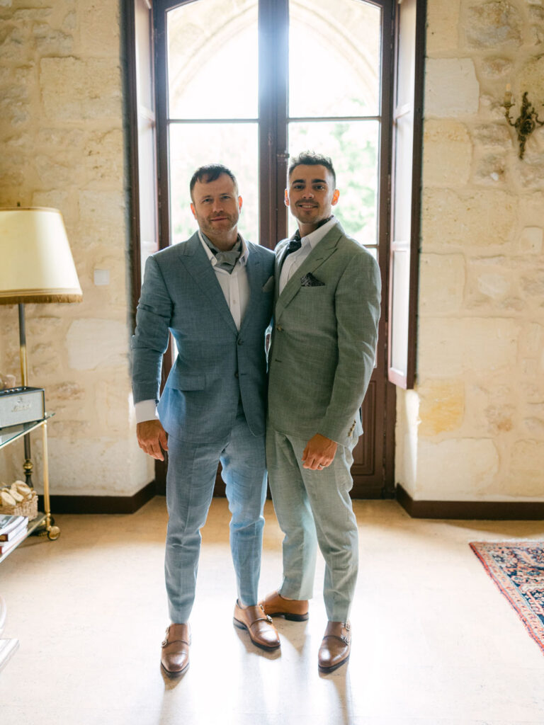 Kevin and Steven dressed in light-colored suits, stand side by side in a well-lit room with stone walls and a window behind ready for their wedding in Francce