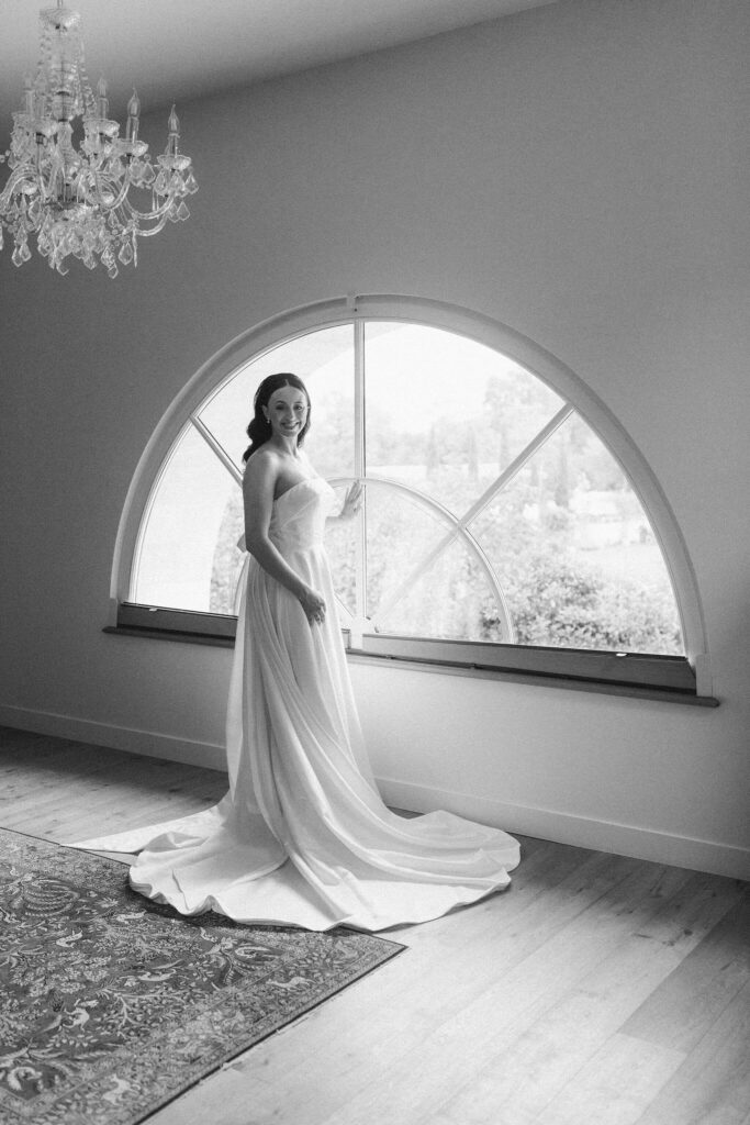 Bride wearing her wedding gown, standing by an arched window