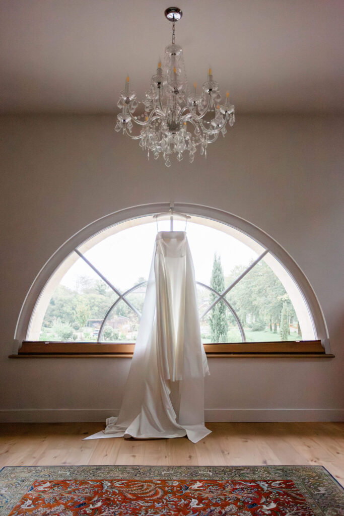 Elegant wedding gown hanging in front of a chandelier and window