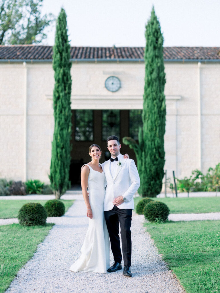 Bride and groom standing together in front of a stone building with cypress trees