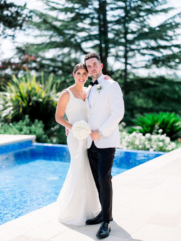 Bride and groom posing by a blue pool with lush greenery in the background