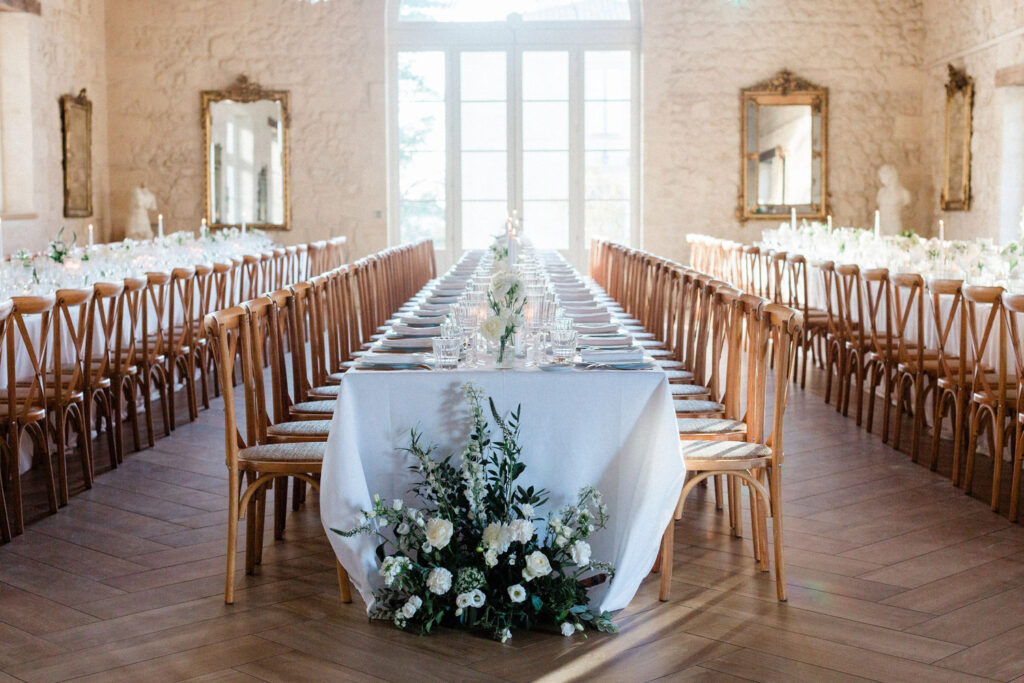 A breathtaking long dining setup with wooden chairs, white florals, and soft natural light filling the elegant chateau space