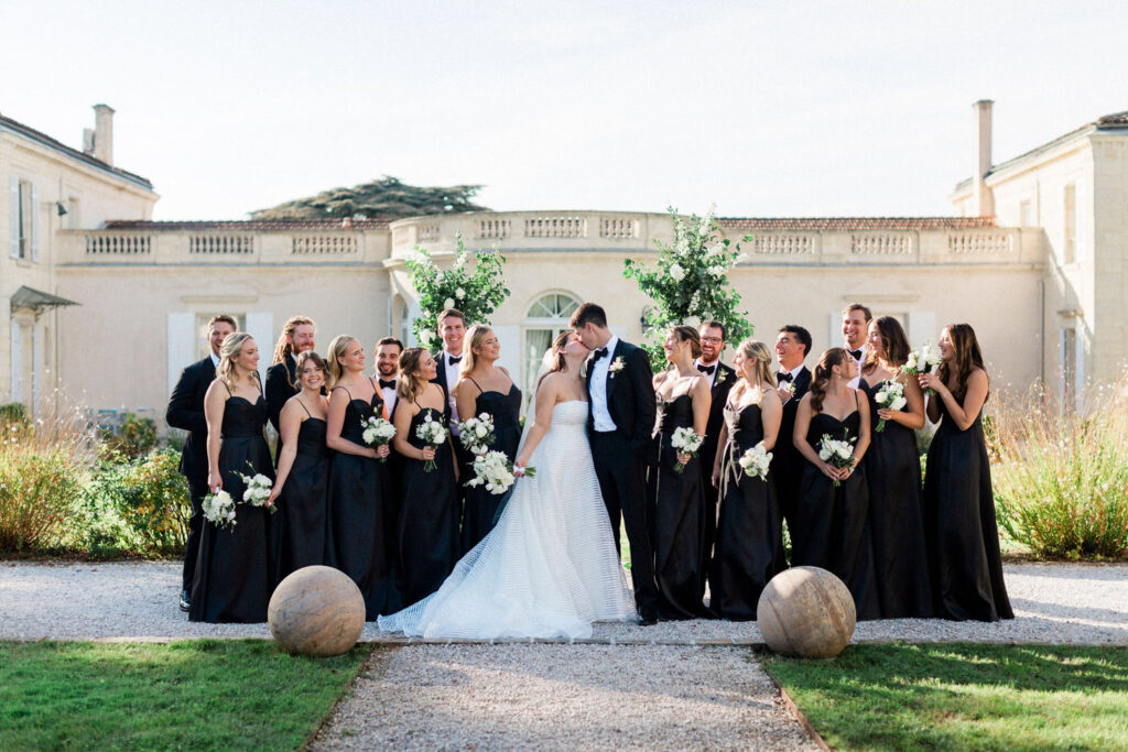 The newlyweds share a kiss surrounded by their bridal party, dressed in timeless black, at their wedding party in Bordeaux