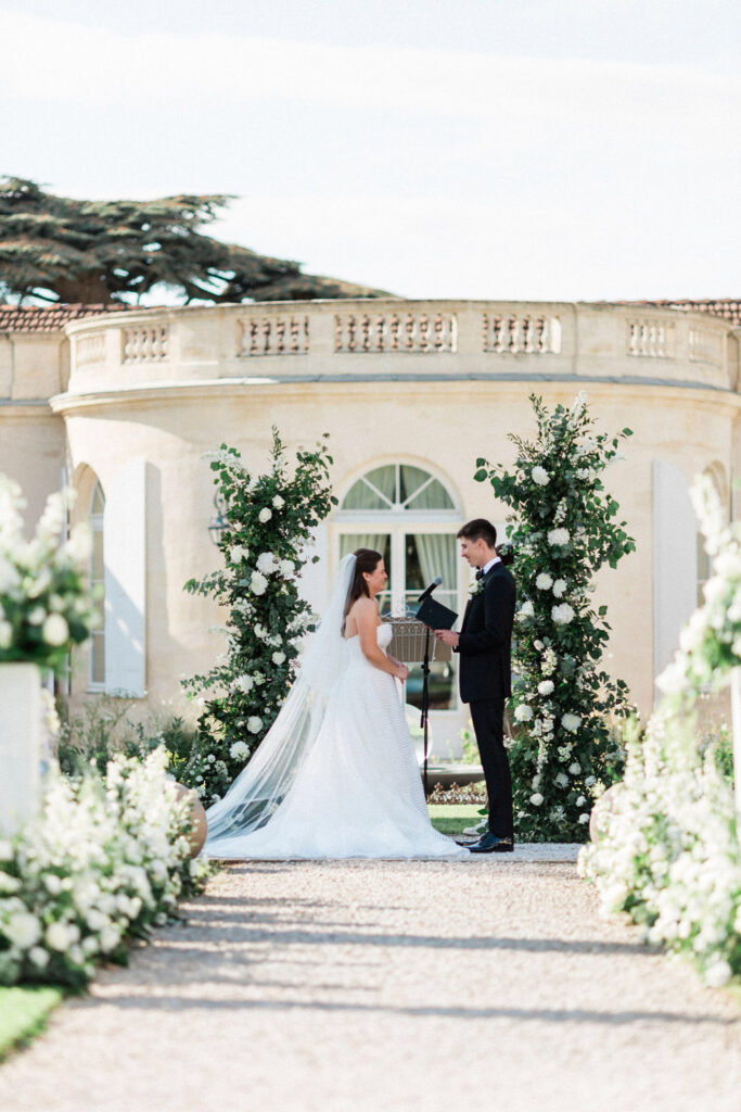 Lauren and Richie exchange vows under a lush white floral arch in a picturesque French château wedding 