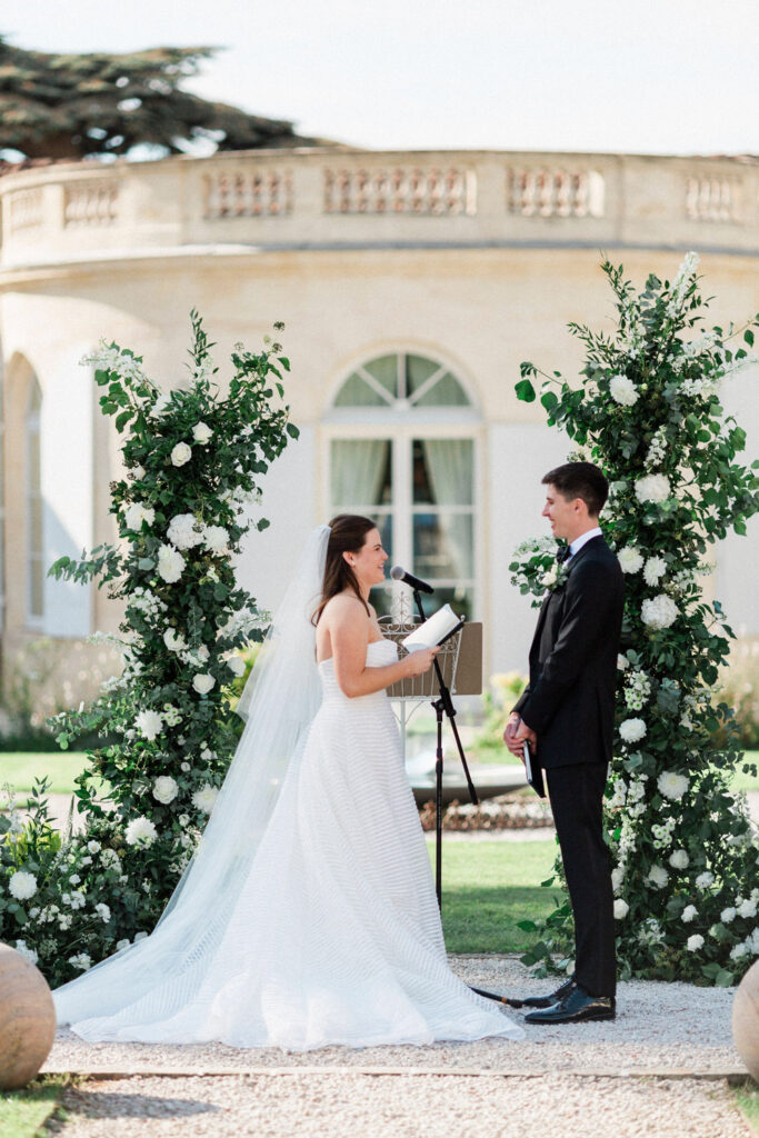 The bride shares her heartfelt vows while the groom smiles, surrounded by greenery and romantic florals.