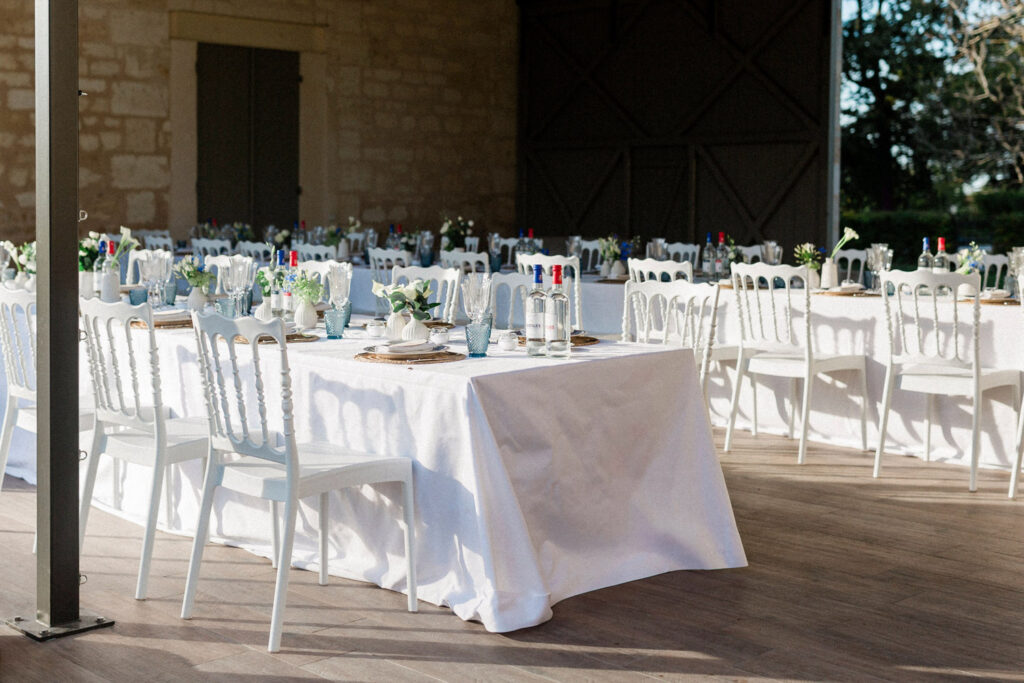 A sunlit wedding dinner setup featuring white chairs, elegant tableware, and fresh florals