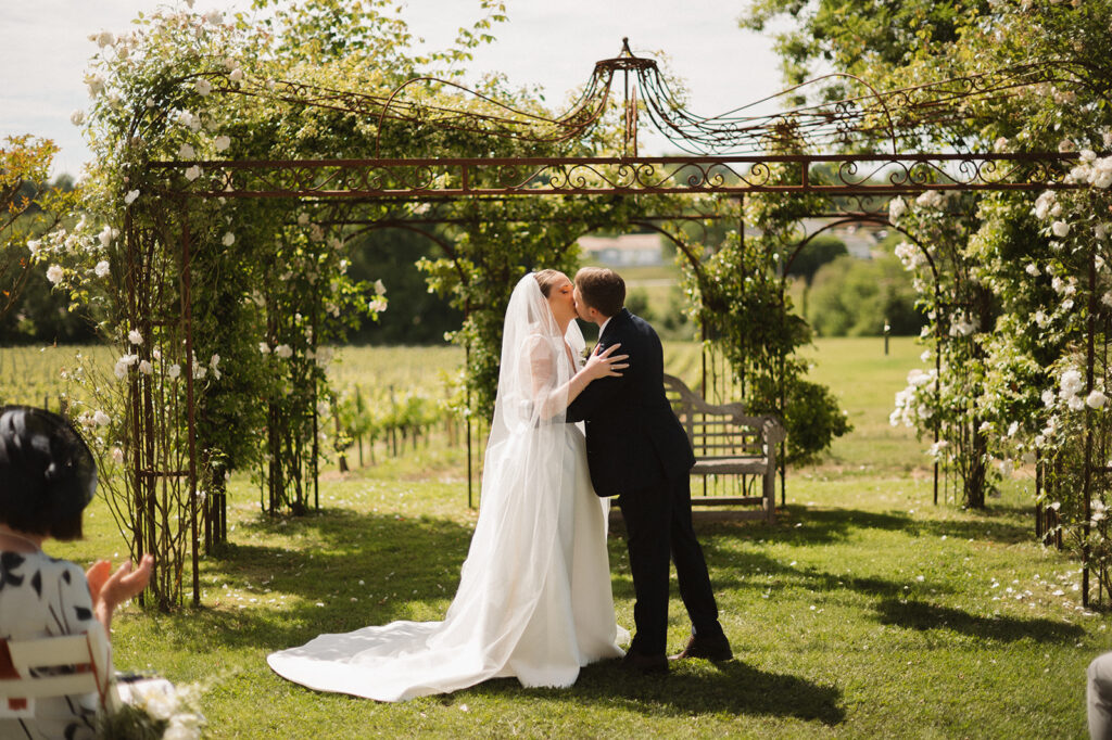 The couple shares their first kiss beneath a floral-covered iron arbor.