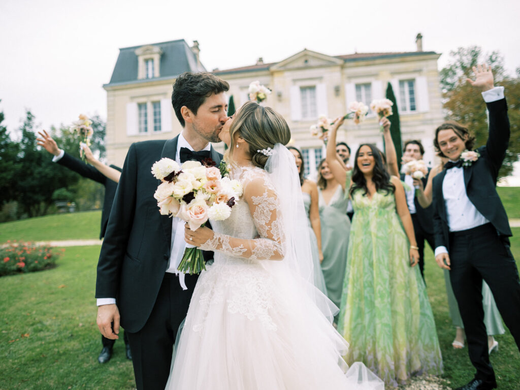 Bride and groom kissing with wedding party celebrating behind them.