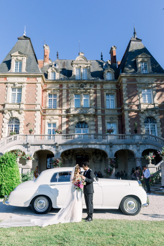 Bride and groom standing beside vintage white car in front of French château