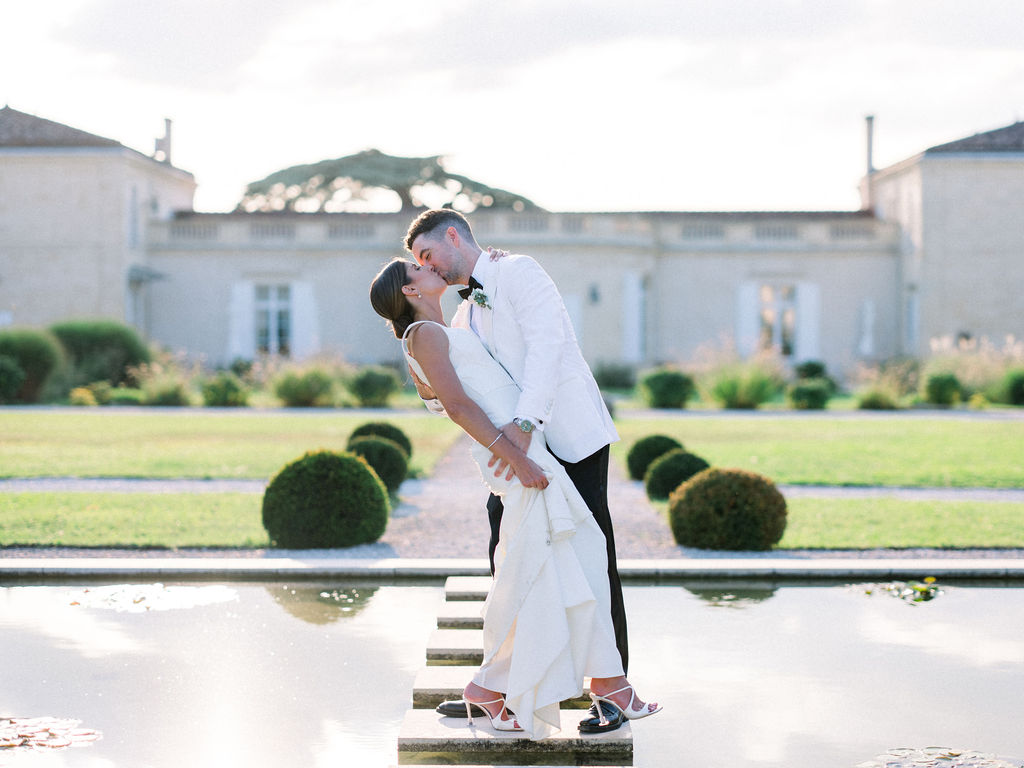 Bride and groom kiss on a stone path over water at a Bordeaux château.