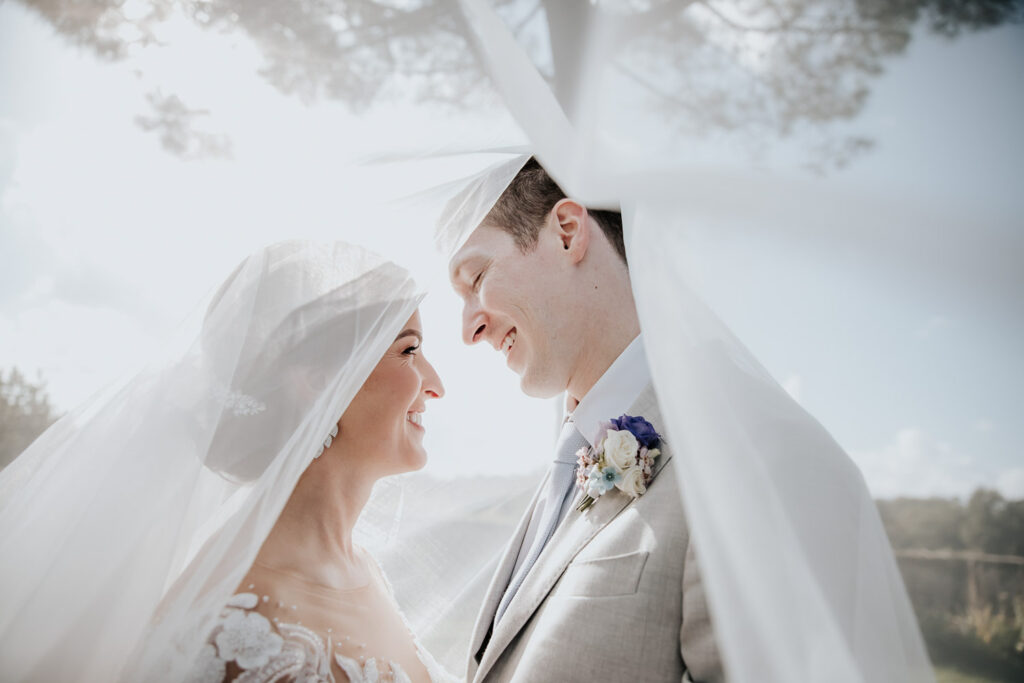 Bride and groom smile under the veil, wrapped in a quiet and intimate moment.