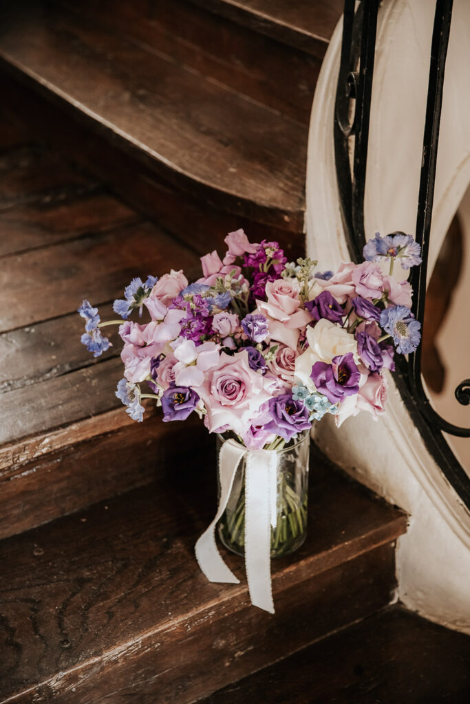 A bouquet of purple and blush florals rests on wooden stairs for a purple wedding inspiration