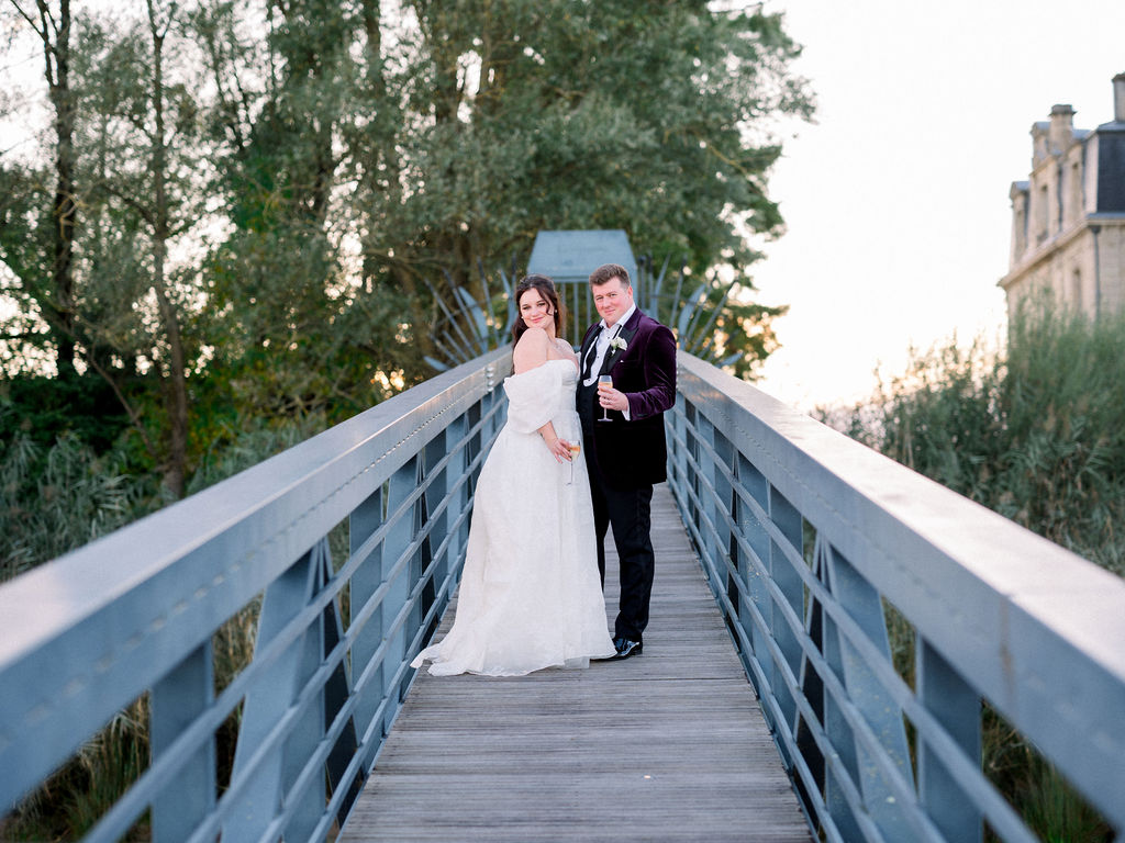 Bride and groom pose with champagne on a wooden bridge near the venue